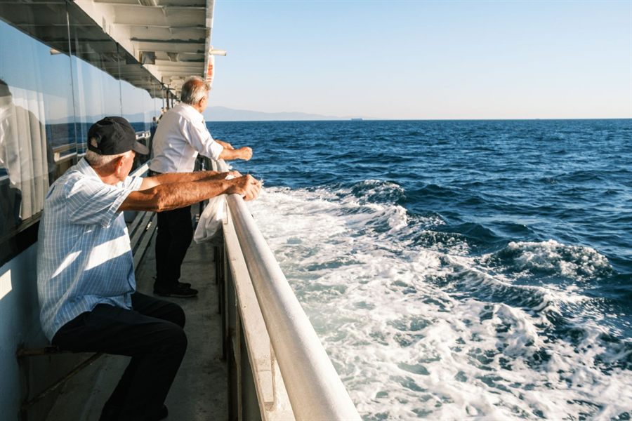 Two older men leaning on a ship rail and watching the wake on a sunny day