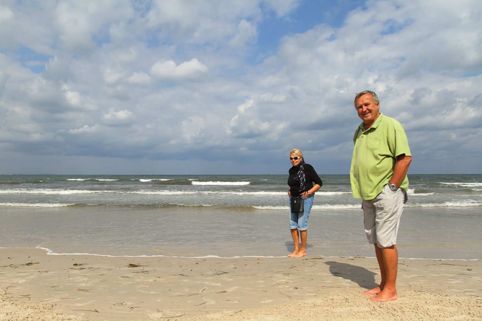 Older couple standing on a sunny beach with waves behind them